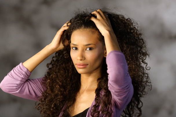 Linda chica afroamericana atractiva tocando su largo y lujoso pelo negro rizado grueso con sus manos mientras se gira para mirar a la cámara - Foto, Imagen