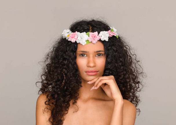 Portret van een mooi meisje uit Santo Domingo met een kapsel van roze en witte bloemen op haar lange krullend zwart haar met de hand tot kin die gepensioneerd naar de camera kijkt - Foto, afbeelding