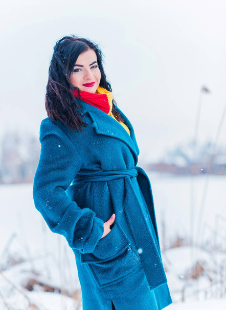 Winter holidays concept. Arabic woman in stylish look at snowy day outside. Woman in blue alpaca coat and red-yellow scarf at winter day outdoors - Foto, afbeelding