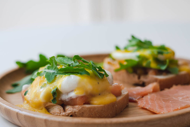 Huevos escalfados Benedict con salsa holandesa, salmón ahumado, rúcula sobre pan tostado. Placa de madera, fondo blanco. - Foto, imagen