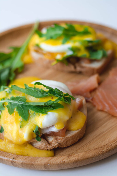 Huevos escalfados Benedict con salsa holandesa, salmón ahumado, rúcula sobre pan tostado. Placa de madera, fondo blanco. - Foto, Imagen