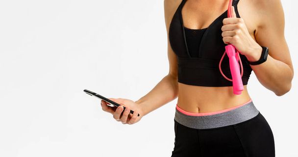 Sporty woman holding smartphone and jump rope on white background. Cardio workout - Photo, Image