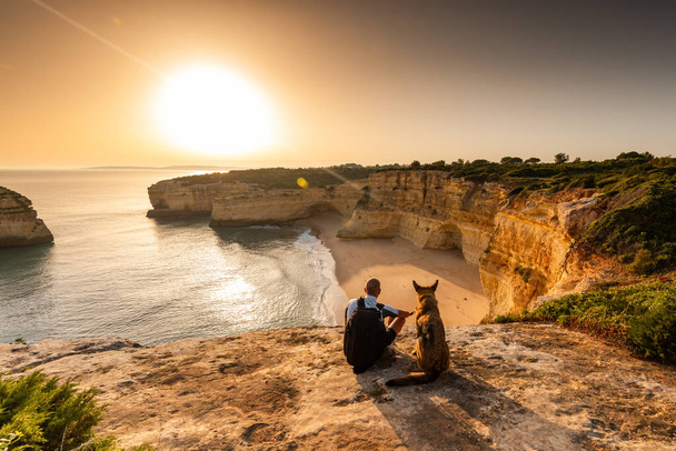 Koncepcja przyjaźni i braterstwa. Mężczyzna i pies patrzący na zachód słońca na klifach morskich w Portugalii. - Zdjęcie, obraz