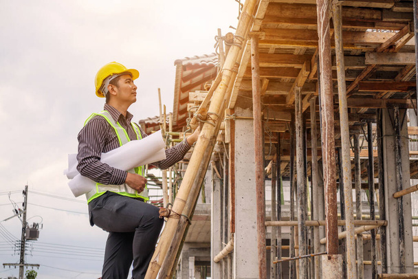 молодой профессиональный инженер-строитель в защитном шлеме и чертежной бумаге под рукой, работающий на лестнице на строительной площадке дома - Фото, изображение
