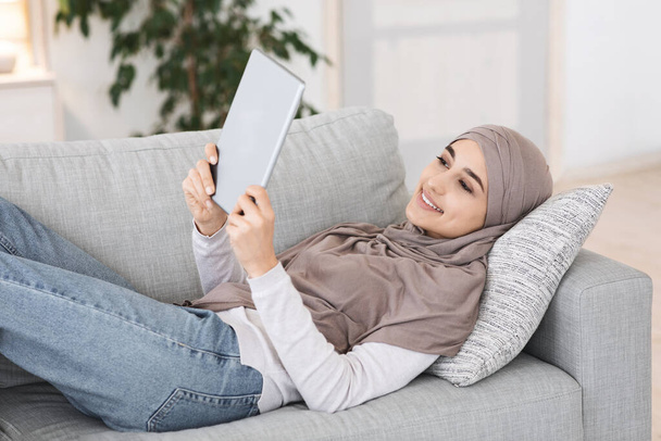 Passage à la maison. Fille musulmane détendue allongée sur le canapé avec tablette numérique - Photo, image