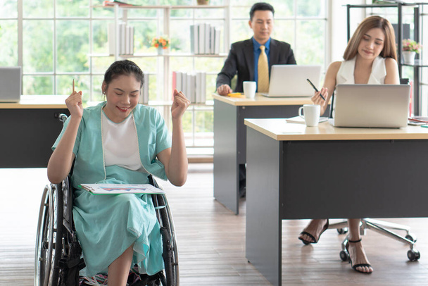 junge asiatische behinderte Frau mit lächelndem Gesicht sitzt im Rollstuhl und benutzt den Computer, um mit ihren Kollegen im Arbeitsbüro das Projekt zu besprechen. Behinderten- und Behindertenkonzept - Foto, Bild