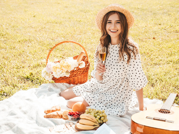 Young beautiful hipster woman in trendy summer sundress and hat. Carefree woman making picnic outside. Positive model sitting on plaid on grass, eating fruits and cheese, drinking champagne - Fotoğraf, Görsel