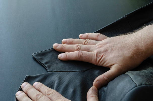 Τα αρσενικά χέρια ισιώνουν το γιακά ενός μαύρου δερμάτινου μπουφάν. Ένα κομμάτι από γνήσιο δερμάτινο ρούχο από κοντά. Επιλεκτική εστίαση. - Φωτογραφία, εικόνα