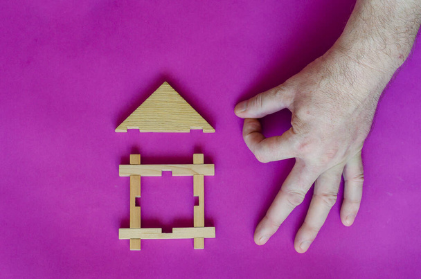 Male hand shares click with sticks on the roof of toy children wooden house on lilac background. Real estate insurance concept. Hand of Caucasian middle-aged man. View from above - Photo, Image