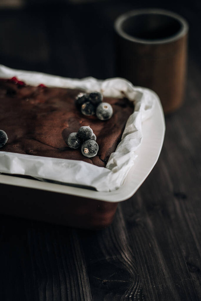 brownie cake garnished with frozen blackcurrant berries in a brown mold with white parchment on a dark background - Foto, immagini