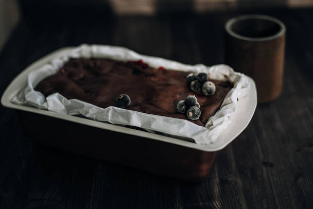 brownie cake garnished with frozen blackcurrant berries in a brown mold with white parchment on a dark background - Fotó, kép
