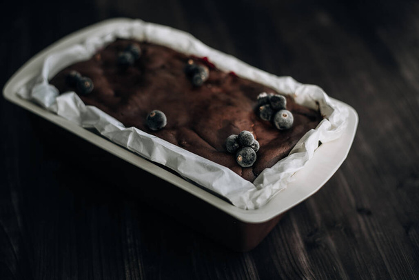 brownie cake garnished with frozen blackcurrant berries in a brown mold with white parchment on a dark background - Photo, image