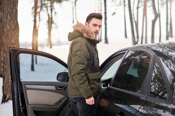 Man stands winter forest road car. Holiday trip. Cold season. Portrait young handsome unshaved driver. Modern hipster guy traveling on car  - Photo, image