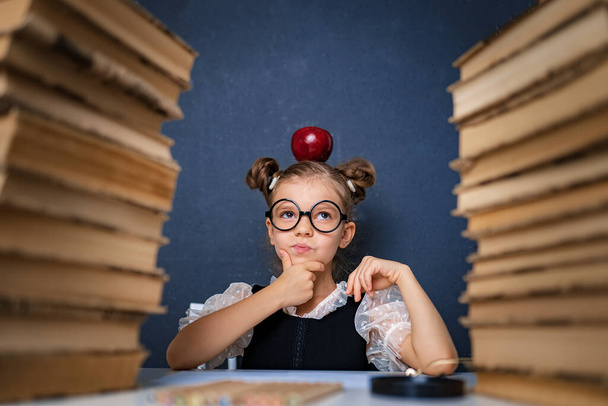Happy smart girl in rounded glasses thoughtfully sitting between two piles of books with red apple on head, pointing finger and look at camera smiling - Foto, afbeelding