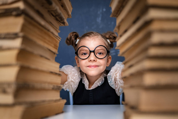Happy smart girl in rounded glasses sitting between two piles of books and look at camera smiling - Zdjęcie, obraz