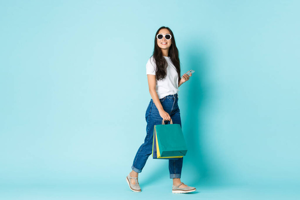 Fashion, beauty and lifestyle concept. Dreamy and happy asian girl enjoying shopping in mall, looking away cheerfully, holding bags and mobile phone over light blue background - Photo, Image