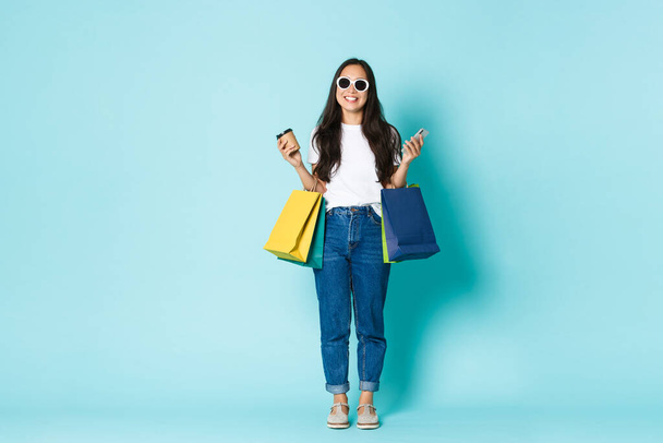 Mode, schoonheid en lifestyle concept. Zorgeloos glimlachende aziatische vrouw genieten van het weekend in het winkelcentrum, het dragen van een zonnebril, het vasthouden van boodschappentassen en het drinken van afhaalkoffie, lichtblauwe achtergrond - Foto, afbeelding