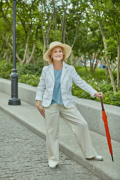 Biała kobieta w wieku około 60 lat stoi w publicznym pakrze w luźnej szacie i z pomarańczowym parasolem. Starsza pani podróżuje, jest aktywna i radosna. - Zdjęcie, obraz
