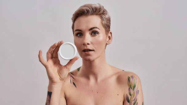 Try it. Portrait of beautiful tattooed woman with pierced nose and short hair holding white plastic jar of cream or body lotion isolated over grey background - Photo, Image