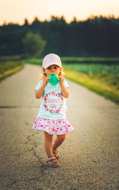 Baby with drinking bottle outdoors walking on a rural road leading to forest. - Photo, Image