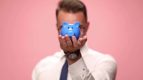 happy elegant businessman in white shirt with glasses holding piggy bank and saving a coin holding fists up and celebrating on pink background - Footage, Video