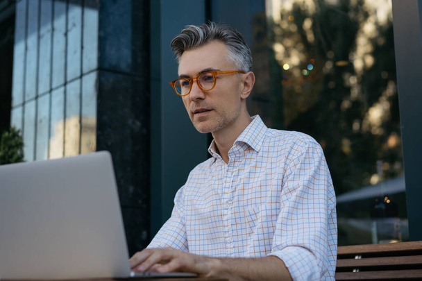 Retrato de desarrollador senior usando computadora portátil, trabajando al aire libre. Hombre de negocios guapo planificación de puesta en marcha, viendo cursos de formación en línea  - Foto, imagen