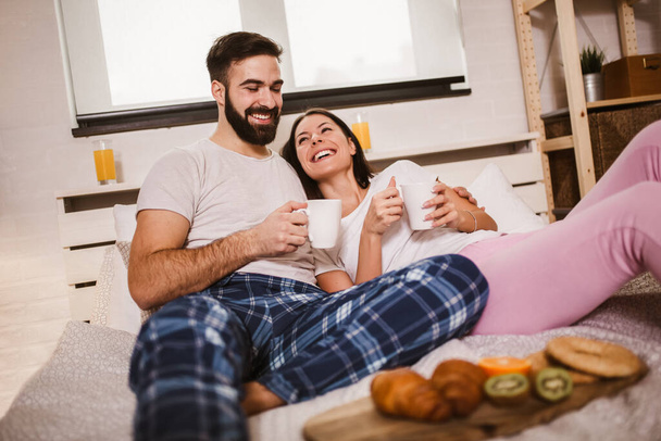 Lächelndes junges Paar im Bett, Kaffee trinkend und einander zuschauend. - Foto, Bild