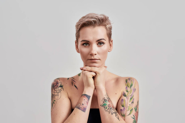 Portrait of half naked tattooed woman with short hair looking at camera, holding chin on hands isolated over light background - 写真・画像