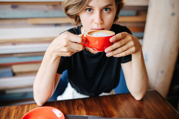 Thoughtful attractive young woman drinking coffee in the corner of cafe, looking at camera with unsettled look. She has dark blond shoulder long hair. Sitting alone. - Fotoğraf, Görsel