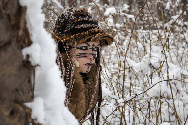Девушка-охотница в одежде и меховой шапке, наблюдает из-за ствола дерева, покрытого снегом, на фоне кустарниковых ветвей - Фото, изображение