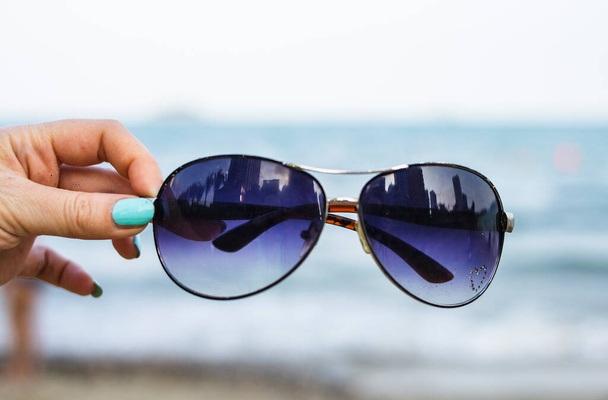 Female hand holds colorful sunglasses against the background of the sky and blue sea. The points reflect the storey buildings of the city. - Fotografie, Obrázek