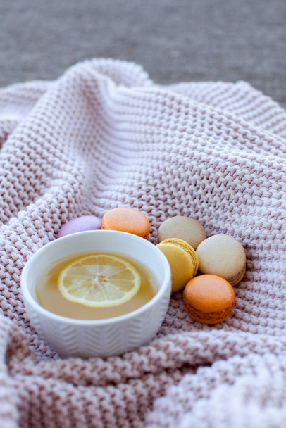 Útulná domácí snídaně s citrónovým čajem, barevnými makaróny na dřevěném podnosu a růžovými kostkami. Koncept sladkostí a dezertů - Fotografie, Obrázek