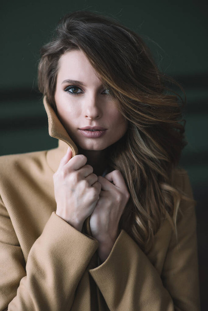 Mooi sexy meisje met krullen en volle lippen in een beige jas in een loft stijl studio. Schoonheid en mode. Zachte selectieve focus. - Foto, afbeelding