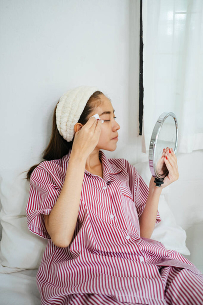 Long hair woman in red pajamas and headband applying toner in front of the mirror. - Foto, Bild