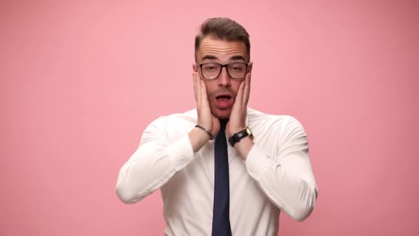 σοκαρισμένος νεαρός άνδρας με λευκό πουκάμισο φορώντας γυαλιά κρατώντας τα χέρια στο πρόσωπο και ανοίγοντας το στόμα, ουρλιάζοντας, κάνοντας γκριμάτσες και προσεύχεται σε ροζ φόντο - Πλάνα, βίντεο
