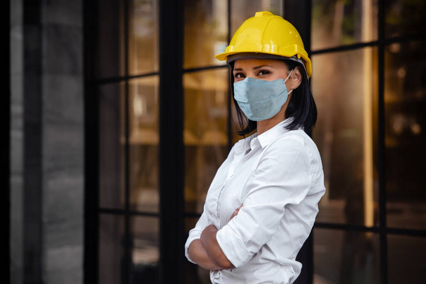 Ritratto di una donna ingegnere edile sicura di sé. Indossare maschera di protezione chirurgica e guardare la macchina fotografica. In piedi di fronte al moderno edificio per uffici - Foto, immagini