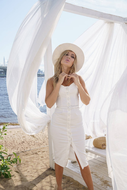Outdoors portrait of beauty blond woman in white dress and white hat. Model posing on beach - Φωτογραφία, εικόνα