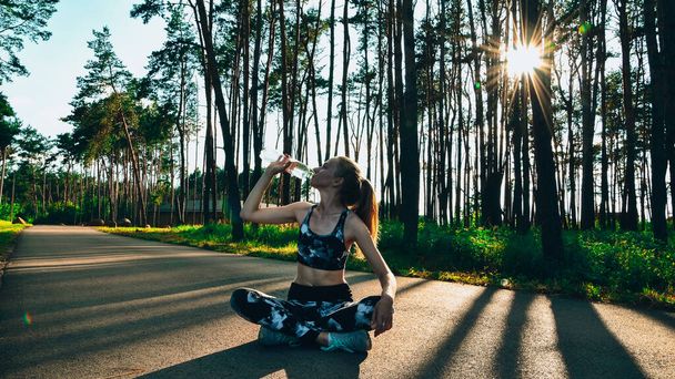 Young woman drinking water after jogging outdoors in forest. Healthy fitness girl sitting in lotus pose and drinking water. Sport outdoor. - Photo, image