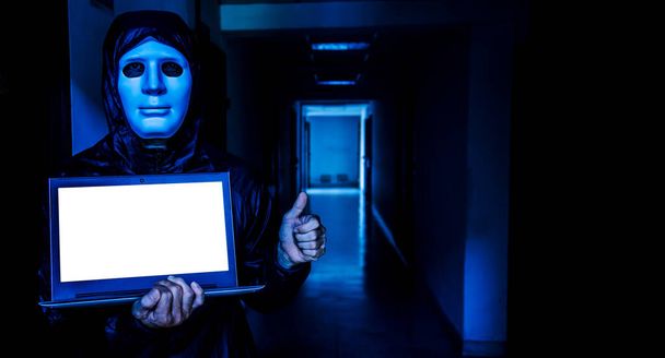 Anonymous computer hacker in white mask and hoodie. Obscured dark face holds a laptop tight in his hands on target room background, Data thief, internet attack, darknet and cyber security concept - Photo, Image