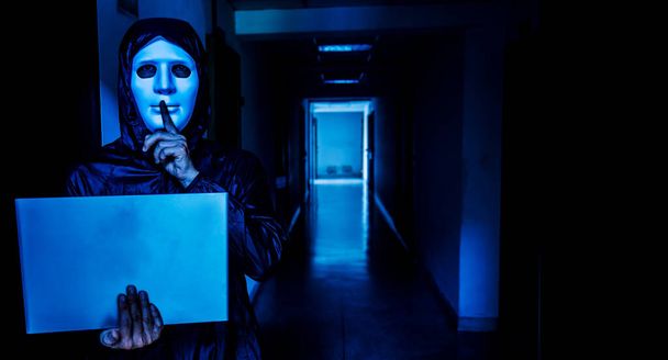 Anonymer Computerhacker in weißer Maske und Kapuzenpulli. Verdunkeltes dunkles Gesicht, Schweigegegeste auf Zielraum-Hintergrund, Datendieb, Internet-Angriff, Darknet und Cyber-Sicherheitskonzept. - Foto, Bild