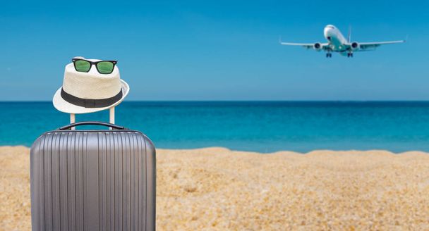 Καλοκαιρινά ταξίδια και σχέδιο με μαύρες αποσκευές βαλίτσα με hipster καπέλο και γυαλιά ηλίου σε τροπική θάλασσα και αμμώδη παραλία, αεροπλάνο και μπλε φόντο ουρανό. Καλοκαιρινές διακοπές ταξίδια έννοια σχεδιασμού - Φωτογραφία, εικόνα