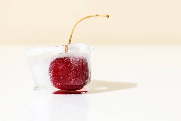 Cherry in ice cube. Concept of shock freezing of berries and frozen food. Preservation of vitamins. Summer harvest. Red ripe sweet cherry with shank on white beige background - Фото, изображение