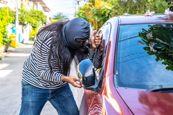 Siyah soygun maskesi takan uzun saçlı erkek hırsız cetvel ve araba hırsızı konseptiyle arabanın camını açmaya çalışıyor.. - Fotoğraf, Görsel