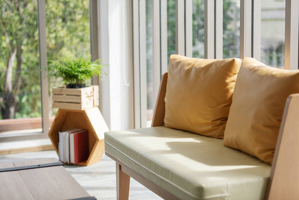 Yellow δερμάτινο μαξιλάρι στον καναπέ κοντά στο παράθυρο στο σαλόνι με φόντο κόκκινο σπίτι στέγη. - Φωτογραφία, εικόνα