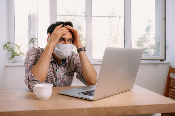 Man in medical mask using laptop on table at home.oung man working on laptop at home at the table in medical mask - 写真・画像