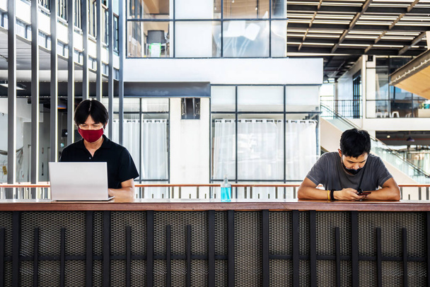2人のアジア人男性がカジュアルなラップトップで事務所で働いている間、ウイルスを保護するためのフェイスマスクを着用し、社会的距離を保ちます。新常態概念. - 写真・画像