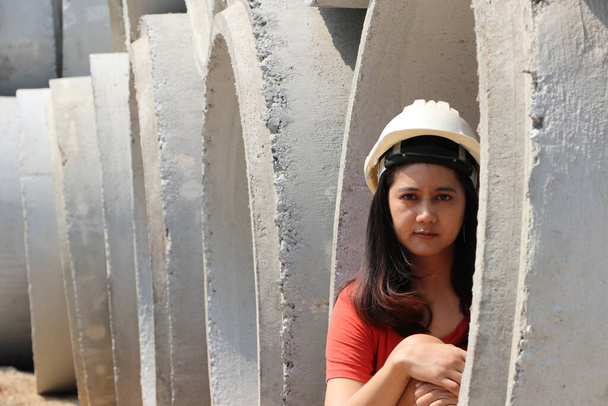 Kadın inşaat mühendisi ya da mimar, kanalizasyon olarak toprağa gömülmeye hazırlanmak için yığılmış büyük beton boruların içinde oturan beyaz miğferi giyer.. - Fotoğraf, Görsel