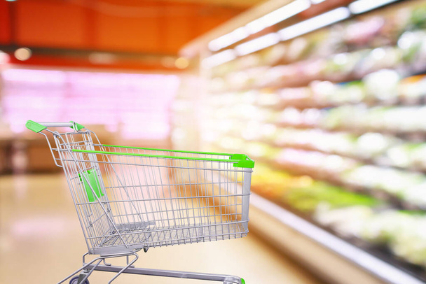 Supermarket aisle with fresh vegetables and fruit shelves interior blur background with empty shopping cart - Photo, Image