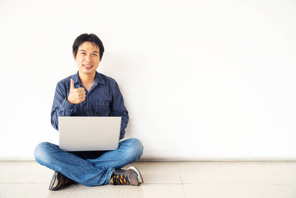 joven hombre asiático sentado en el suelo con el uso de computadora portátil feliz con gran sonrisa haciendo ok signo, pulgar hacia arriba con los dedos, excelente signo - Foto, Imagen