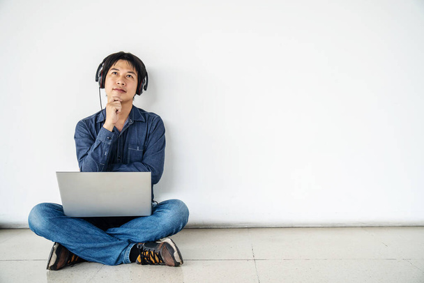 Joven asiático sentado en el suelo con el uso de ordenador portátil y escuchar los datos en sentir dudas y confundido o pensando en una idea posando aislado sobre la pared blanca, espacio de copia - Foto, Imagen